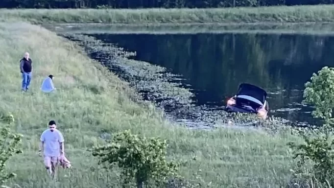 A photo of people on the shore of a pond with a car half submerged in the inky water.